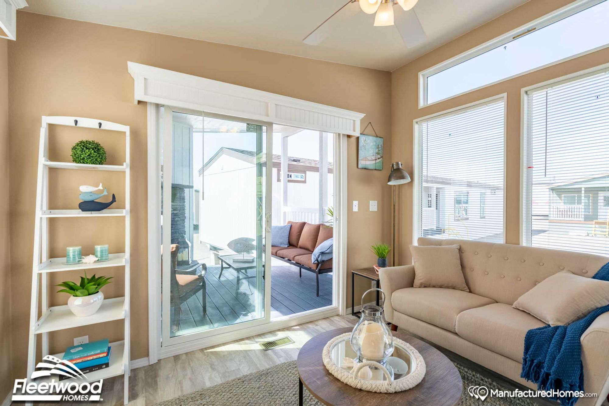 Cozy living room with beige walls, a ceiling fan, and sliding glass doors opening to a patio with outdoor seating. The space features a round coffee table with decor, a ladder shelf, framed artwork, and natural light from large windows.