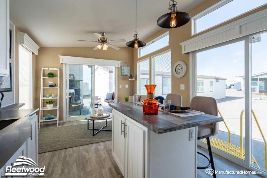 Bright dining and kitchen area with large windows, sliding glass doors, a breakfast bar with seating, and modern pendant lights. An orange glass vase decorates the countertop, while a ceiling fan and wall clock add to the cozy space.