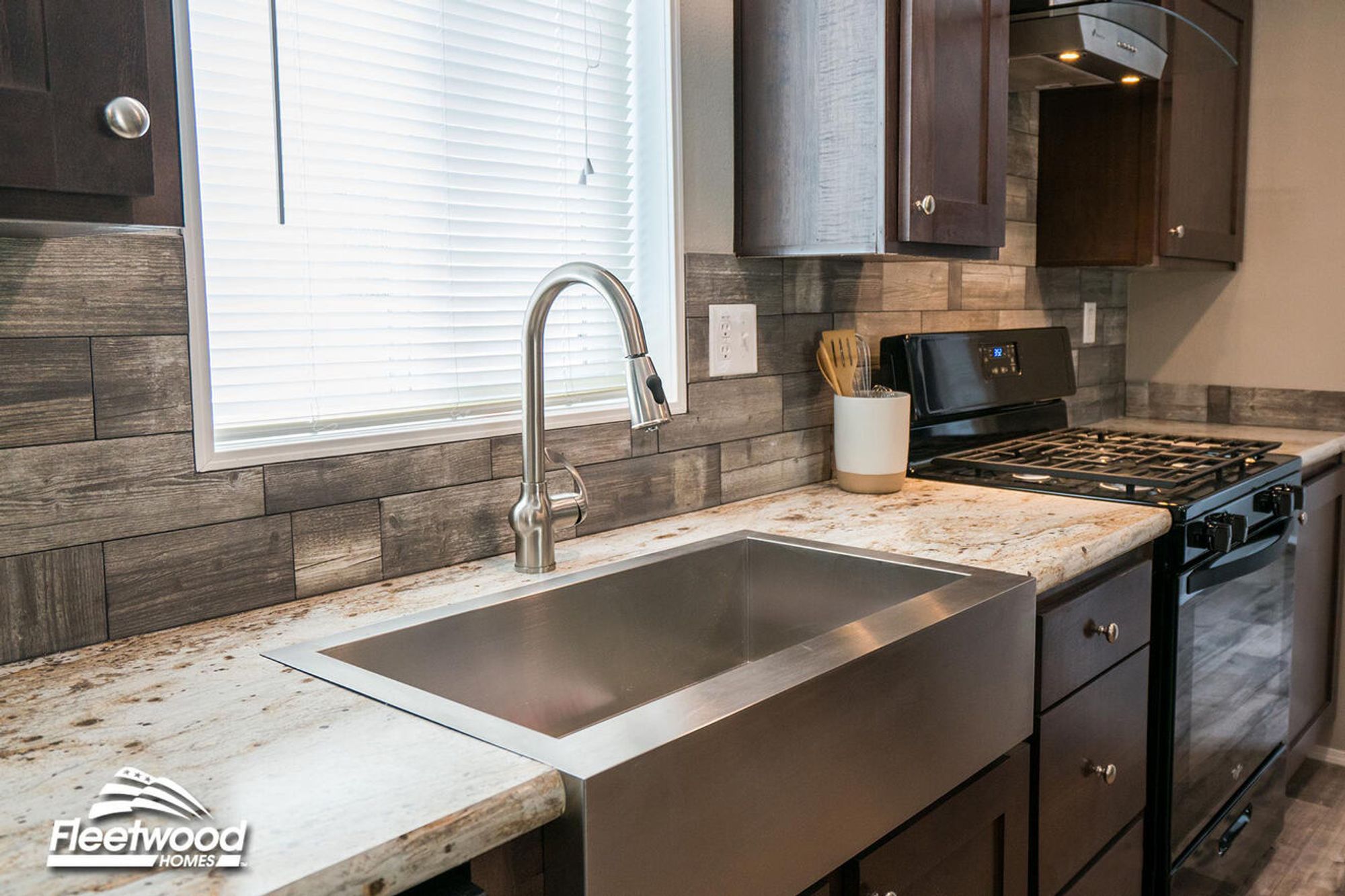 Close-up of a stainless steel farmhouse sink under a window, set in a granite countertop with a gray wood-textured tile backsplash and dark wood cabinetry.