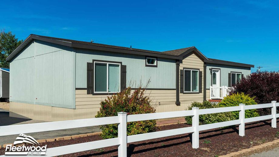 Manufactured home with beige and light blue siding, dark trim, front porch entry, and white fencing with landscaped shrubs.