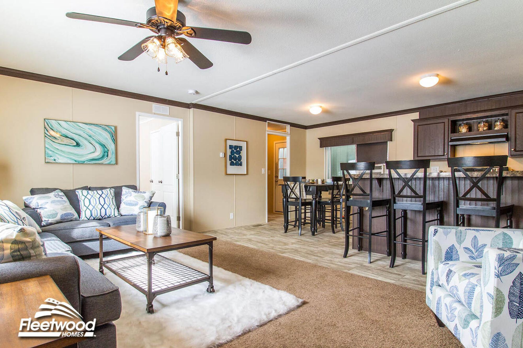 Open-concept kitchen and living room with a breakfast bar and barstools, modern appliances, carpeted seating area with sofas and an accent chair, ceiling fan, and a wooden coffee table on a rug.