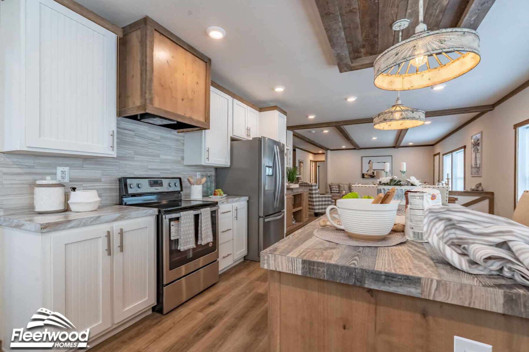 Open-concept kitchen and dining area with a rustic wood island, pendant lighting, wood-look flooring, and large windows, flowing into a living room with a sectional sofa.