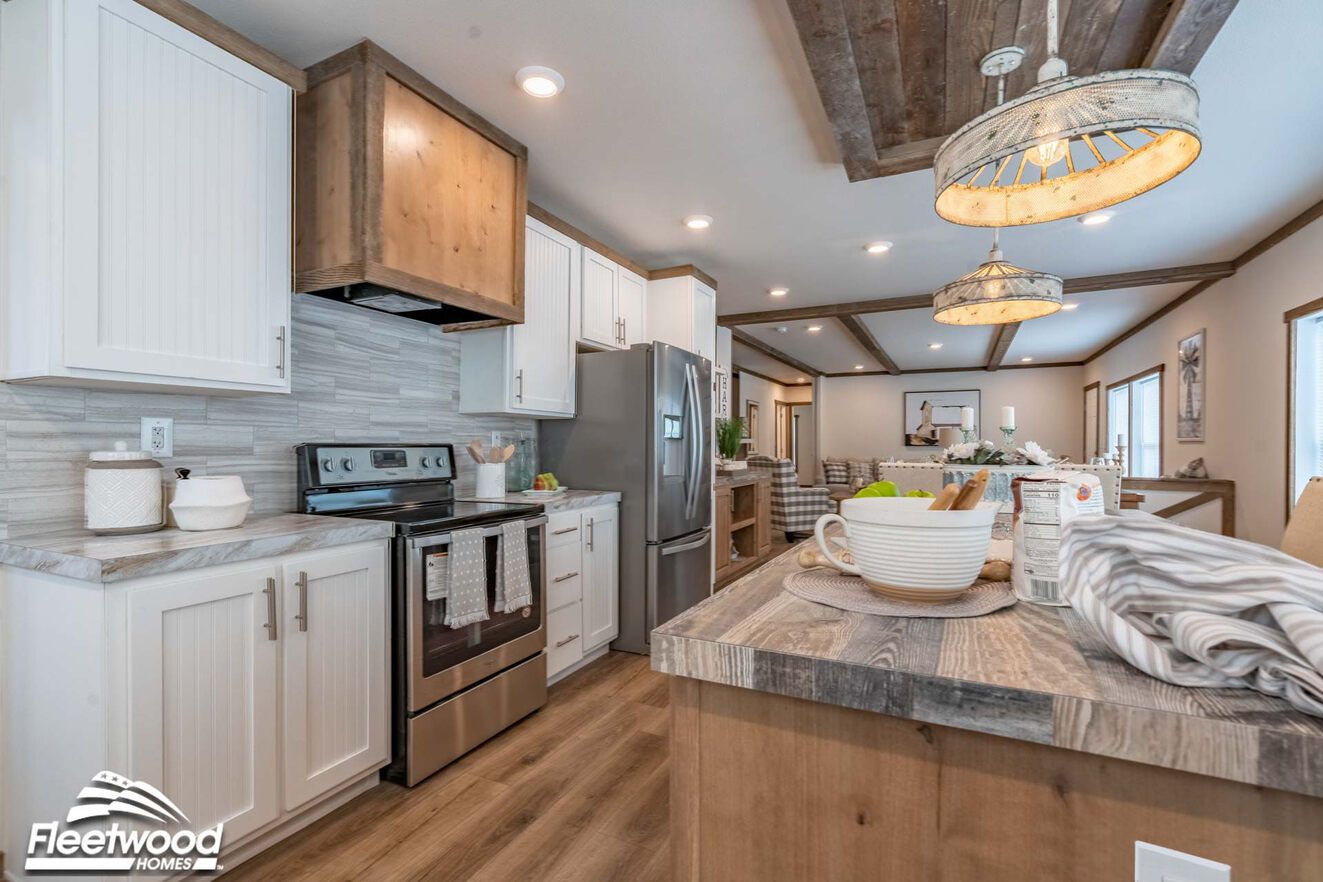 Open-concept kitchen and dining area with a rustic wood island, pendant lighting, wood-look flooring, and large windows, flowing into a living room with a sectional sofa.