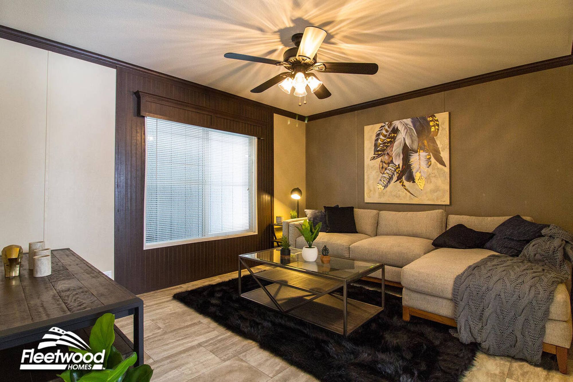 Living room with sectional sofa, glass coffee table on a black rug, ceiling fan, and large window with blinds.