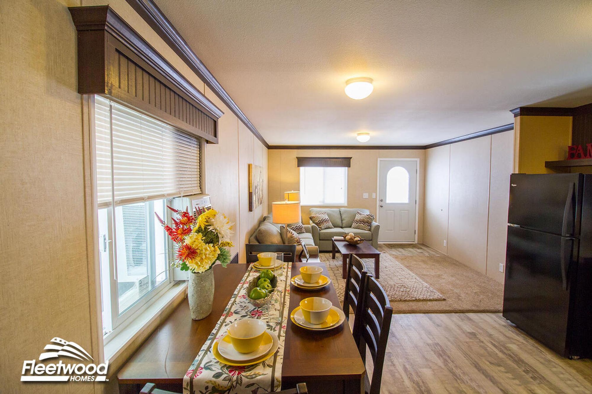 Dining table set with yellow bowls and plates beside a window with blinds, decorated with a floral runner and vase of flowers, looking into a living room with a sofa, armchair, coffee table, and front door; part of the kitchen with a black refrigerator is visible on the right.