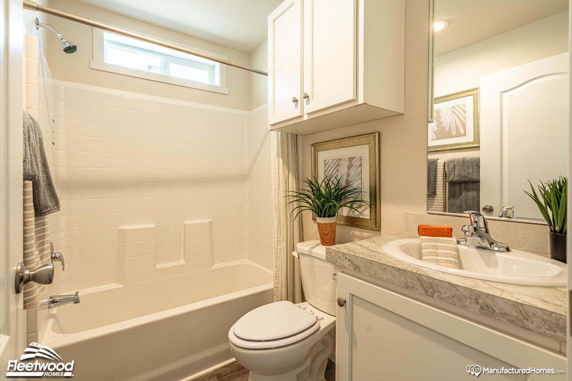 Bright bathroom with a white shower-tub combo, striped shower curtain, and window above. Gray and striped towels hang nearby, with white flowers placed on the toilet tank lid.