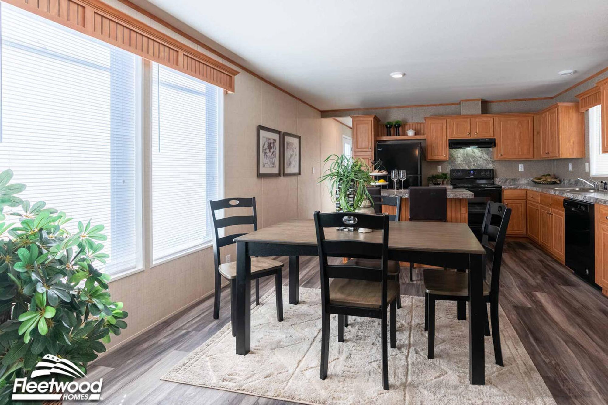 Dining area with a wooden table and black chairs beside large windows, opening into a kitchen with wood cabinets and black appliances.