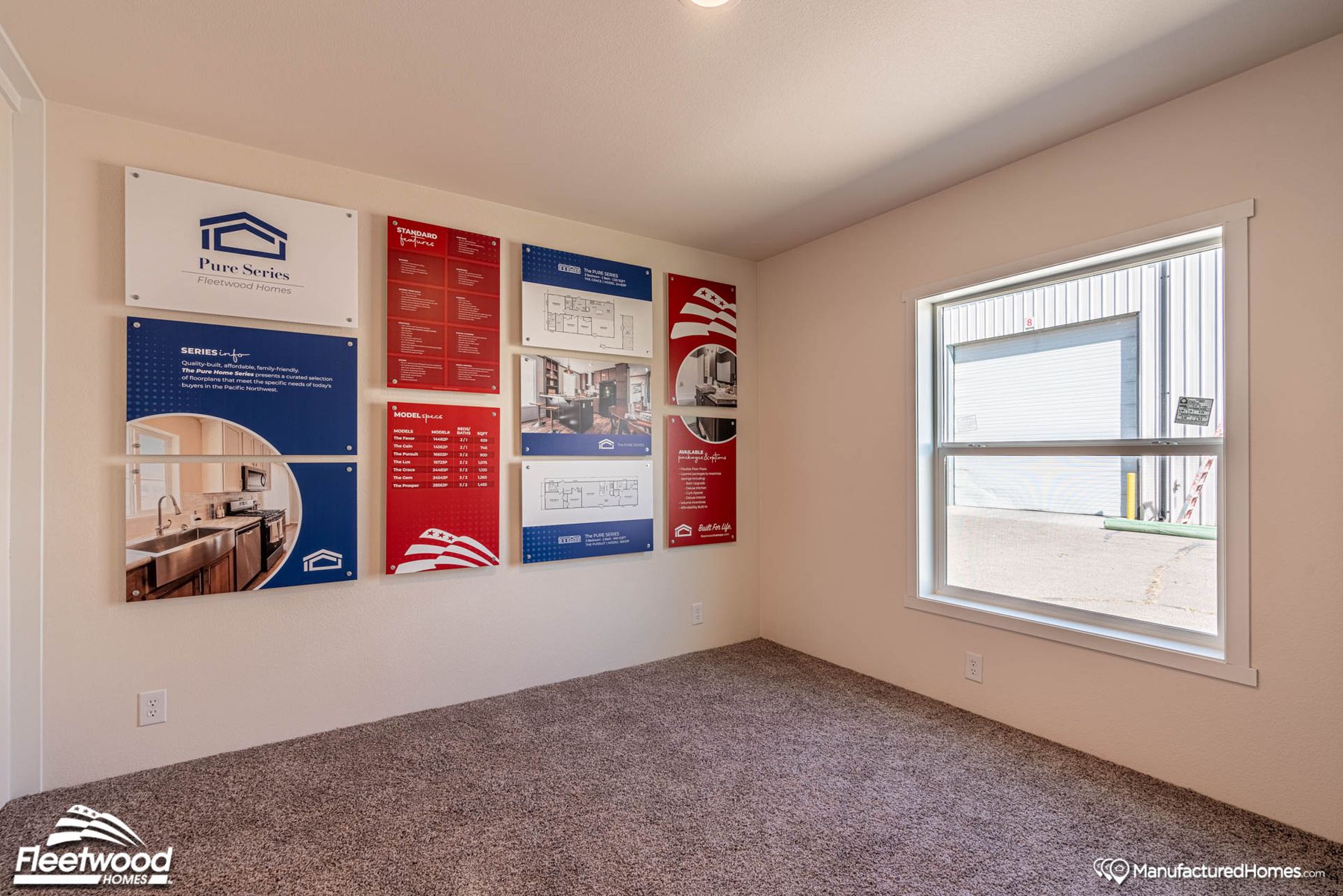 Room with beige walls and carpeted flooring, featuring Cavco Idaho product info boards on the wall. A large window provides natural light and a view of a metal building outside.