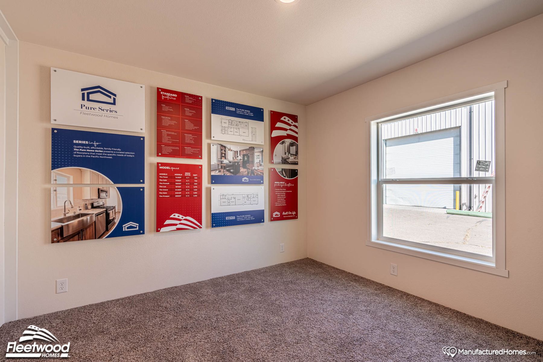 Room with beige walls and carpeted flooring, featuring Cavco Idaho product info boards on the wall. A large window provides natural light and a view of a metal building outside.