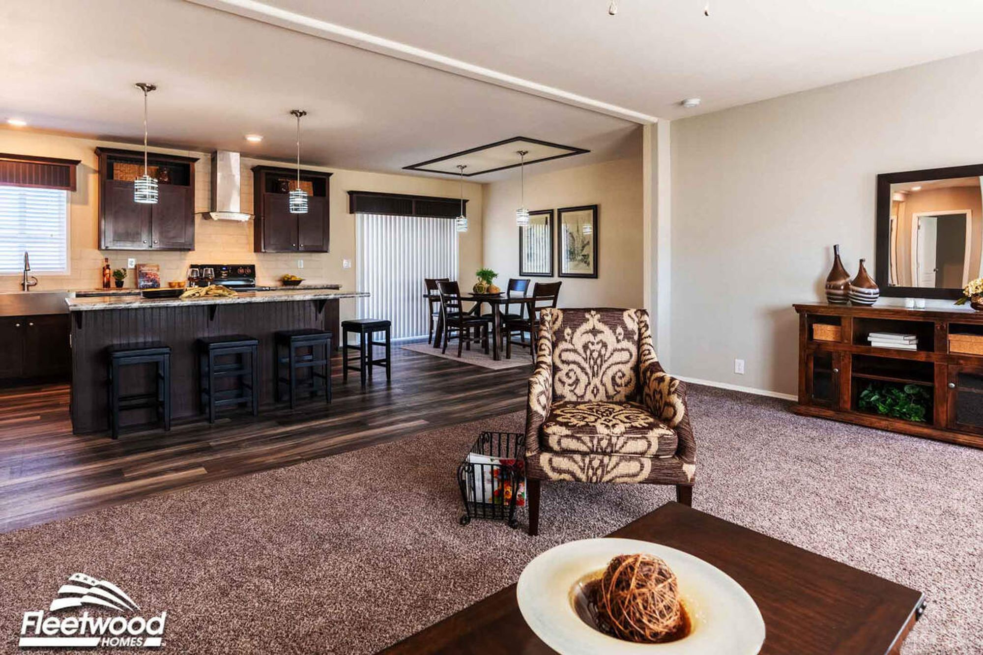 Open living room with brown sofas, patterned accent chair, and view of a modern kitchen with island seating and dark cabinetry.