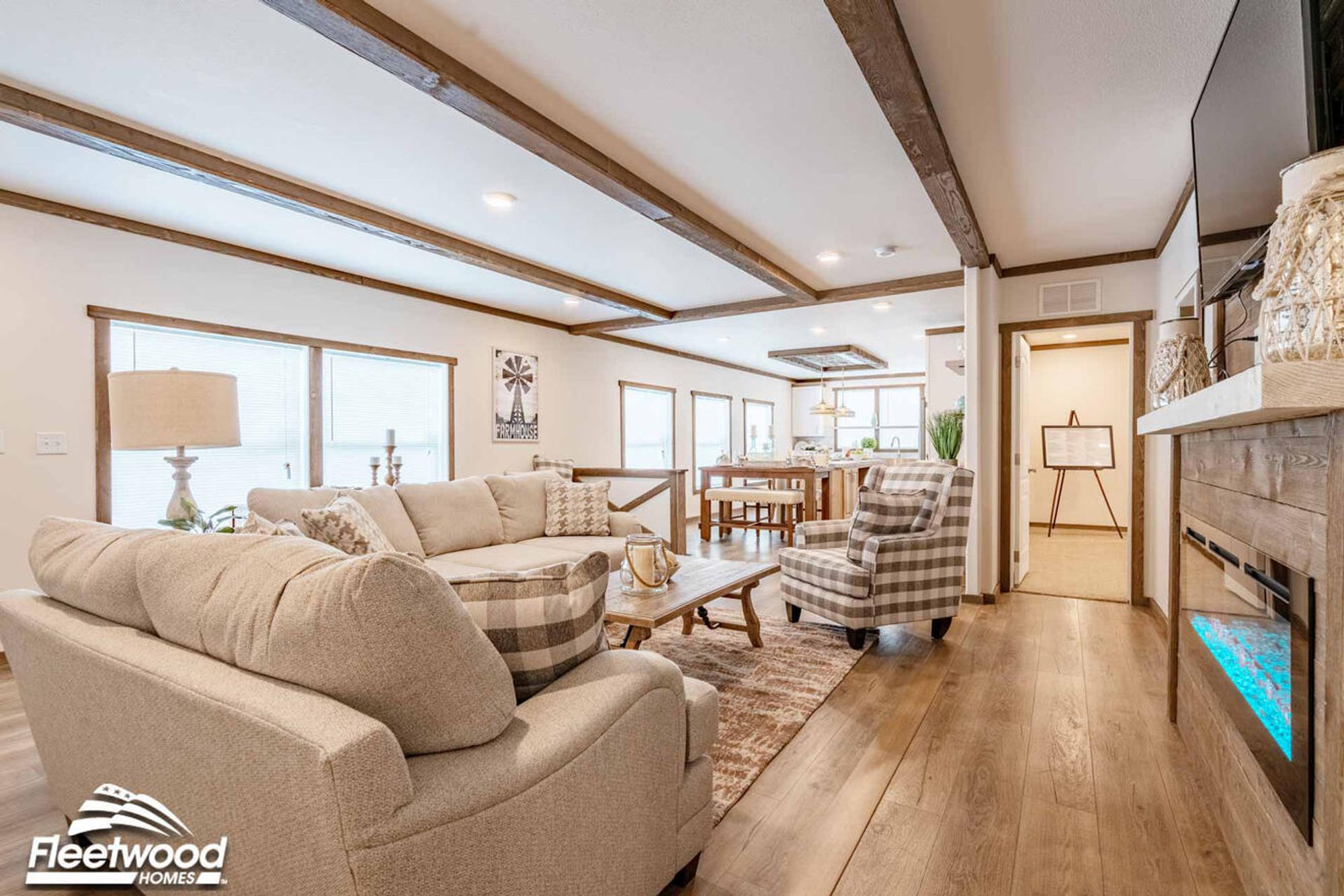 Cozy living room with wood-beam ceiling accents, neutral-toned sofas with patterned pillows, a plaid armchair, a rustic coffee table, and a wood-paneled fireplace wall with a mounted TV.