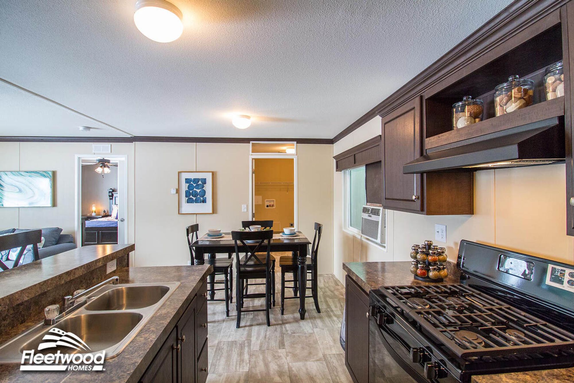 Kitchen with dark cabinets, double sink, and stove, opening to a dining area with a table and chairs; living room and bedroom visible in the background.