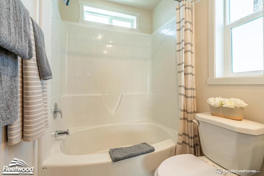 Bright bathroom with a white shower-tub combo, striped shower curtain, and window above. Gray and striped towels hang nearby, with white flowers placed on the toilet tank lid.