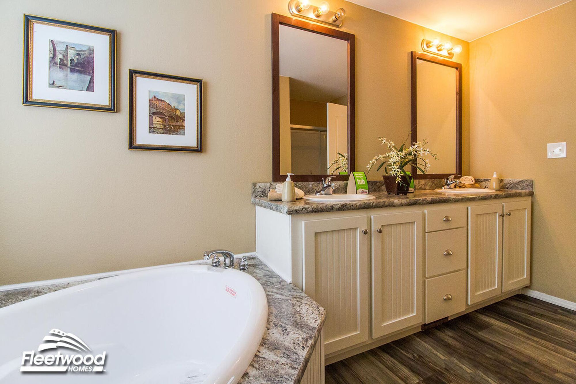 Warm-toned bathroom with a corner soaking tub, double-sink vanity with granite countertops, two mirrors, and wall-mounted artwork above the tub.