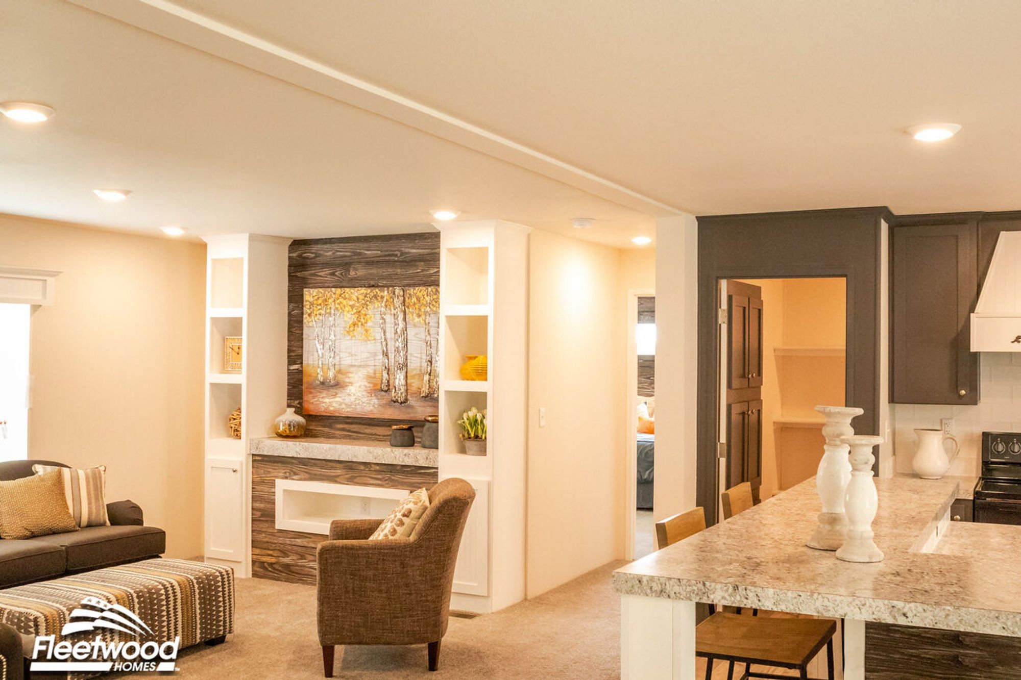 Open-concept living space with neutral tones, built-in entertainment center featuring a birch tree painting, and a view into a cozy bedroom and kitchen with a granite peninsula.