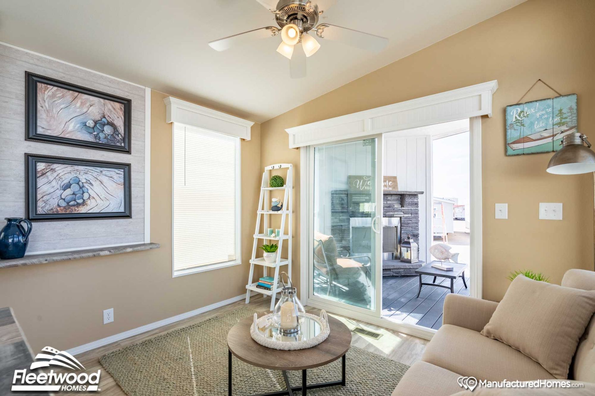 Cozy living room with beige walls, a ceiling fan, and sliding glass doors opening to a patio with outdoor seating. The space features a round coffee table with decor, a ladder shelf, framed artwork, and natural light from large windows.