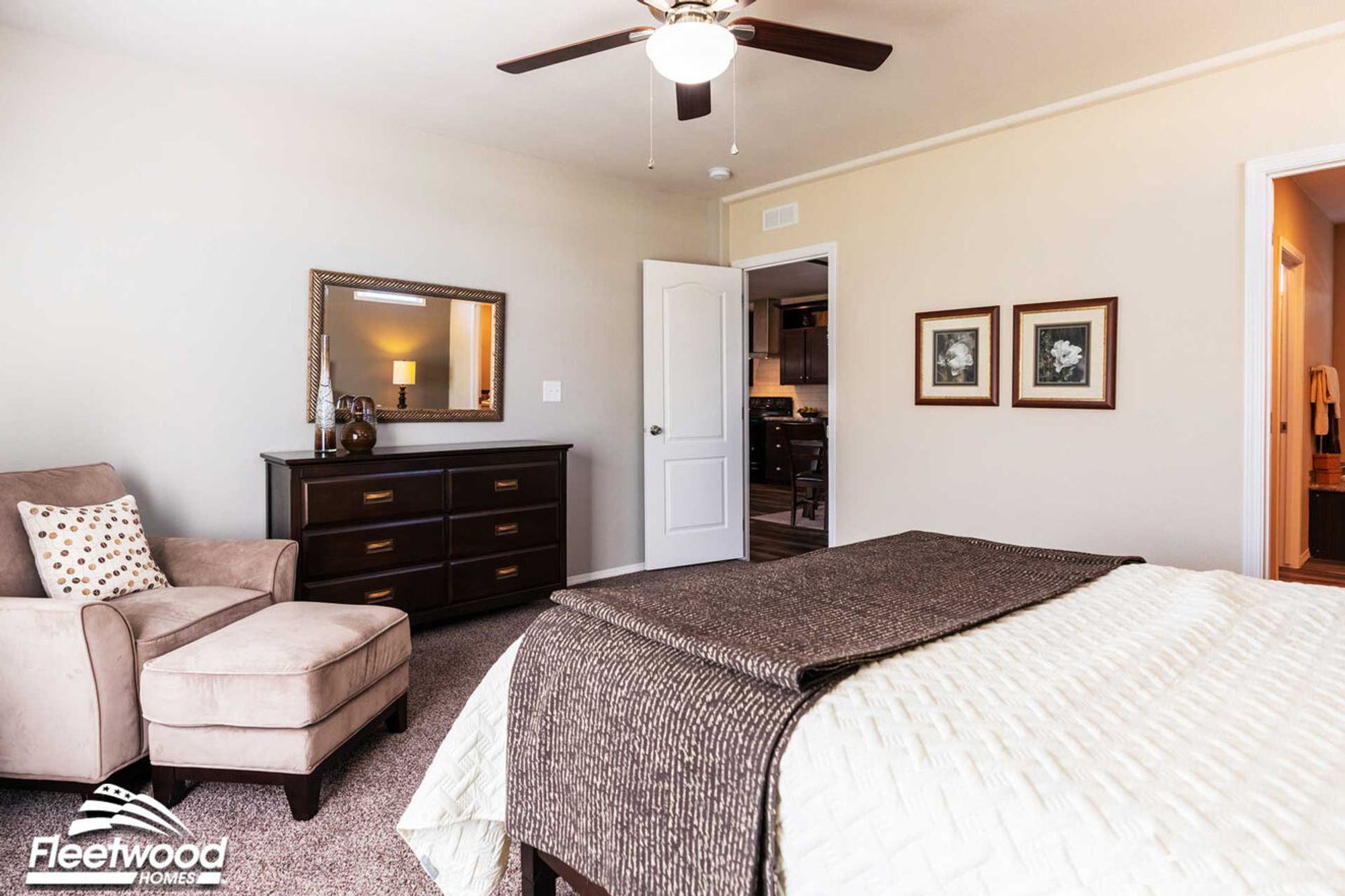 Bedroom with neutral decor, featuring a bed with white and brown bedding, dark wood dresser, beige armchair, and ceiling fan.