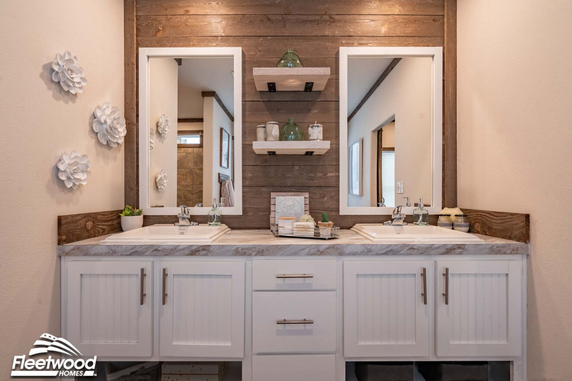 Bathroom vanity with dual sinks, marble-style countertops, white cabinetry, two framed mirrors, wood accent wall, and floating shelves with decorative items.