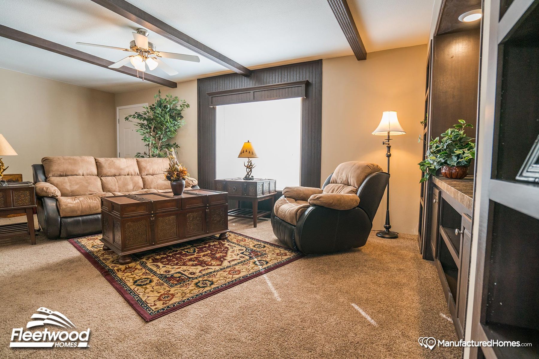 Cozy living room in a manufactured home, featuring beige recliner seating, a dark wood coffee table, and decorative plants on plush carpet with a patterned area rug.