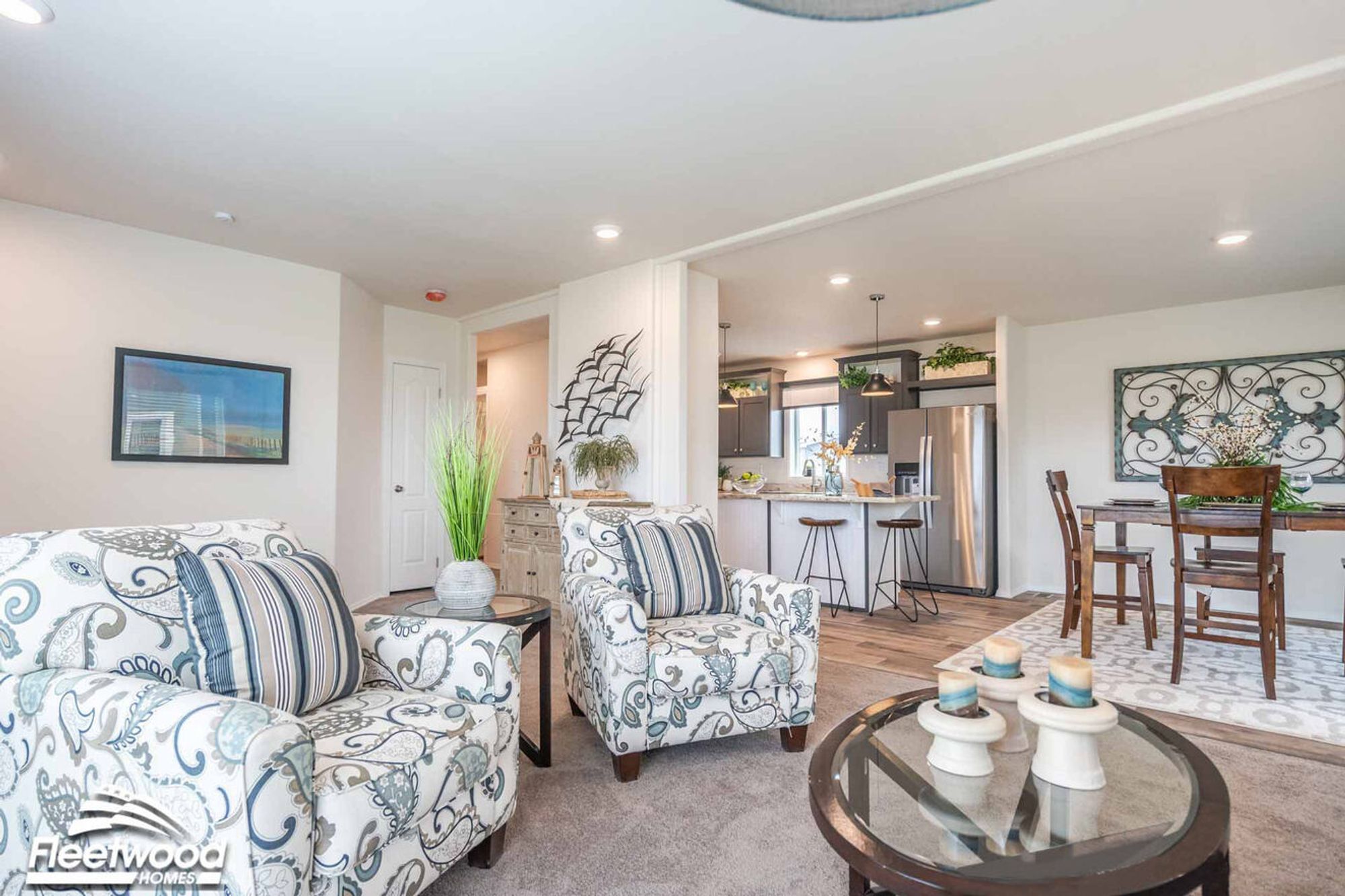 Open living space with patterned armchairs, glass coffee table, and view into a modern kitchen with bar seating and dining area.