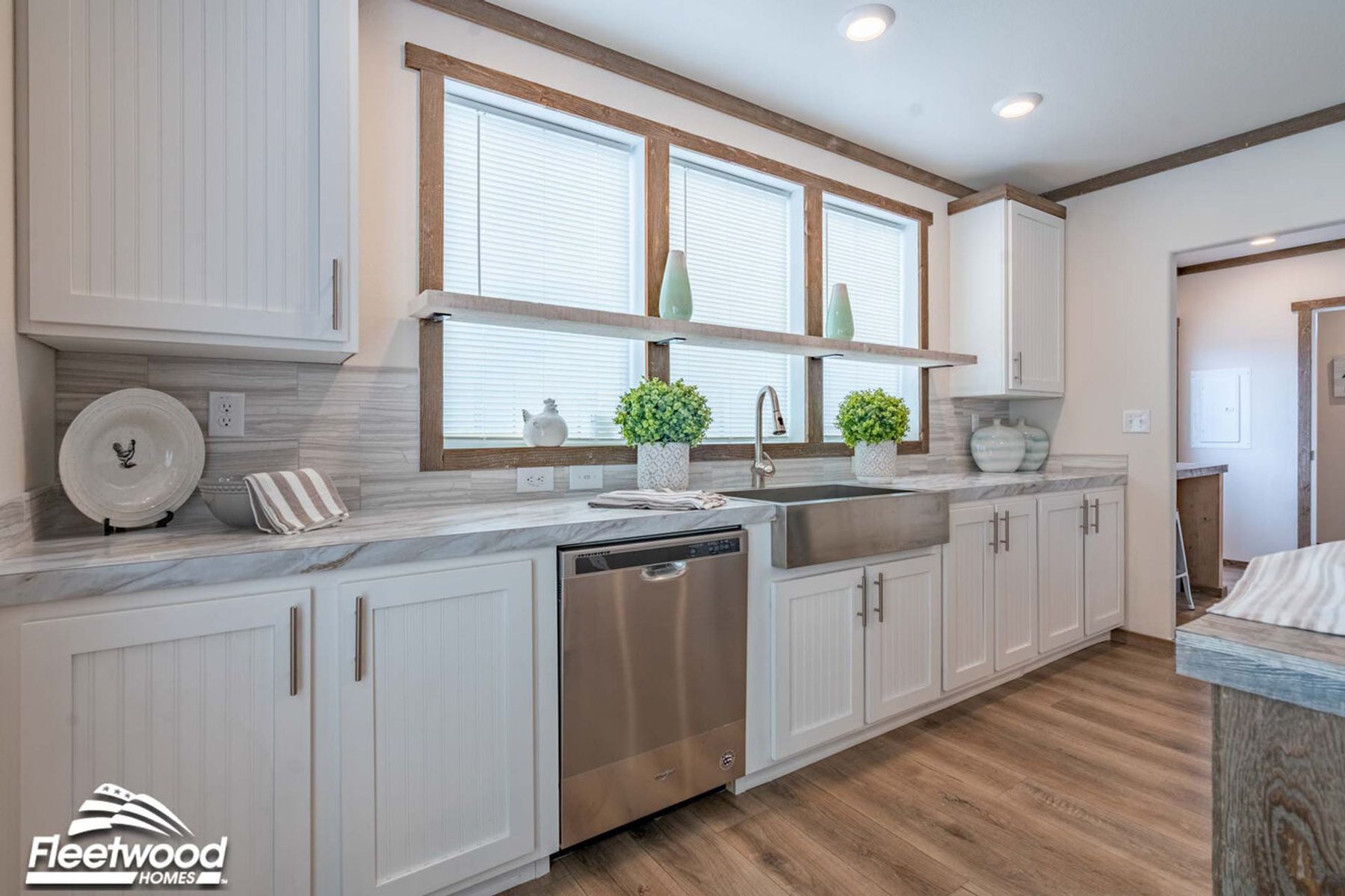 Modern kitchen with white cabinetry, stainless steel farmhouse sink, dishwasher, and wood-look flooring, featuring a long counter under three large windows with a decorative shelf.
