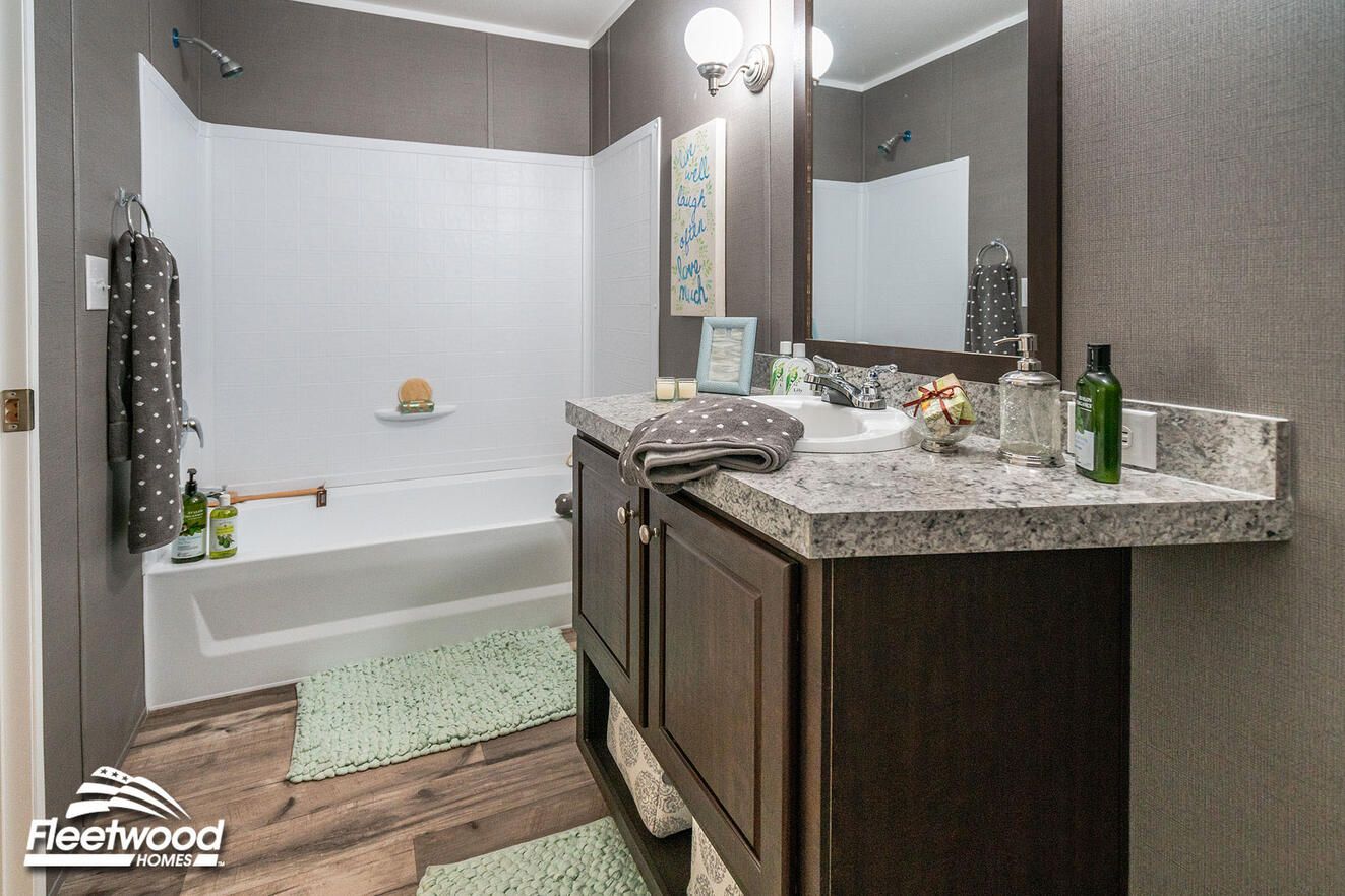 Bathroom with a granite-style vanity, dark wood cabinets, a white sink, and a bathtub with a shower combo. Green rugs add contrast to the wood-style flooring.