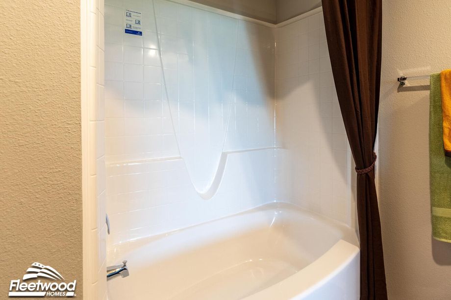 Clean white bathtub with built-in shower, brown shower curtain pulled to the side, and green and orange towels hanging nearby.