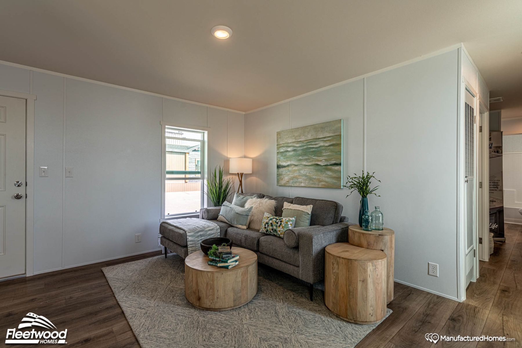 Bright and modern living room with a gray sectional sofa, decorative pillows, round wood coffee tables, and soft natural light from two windows, styled with coastal-themed artwork and plants.