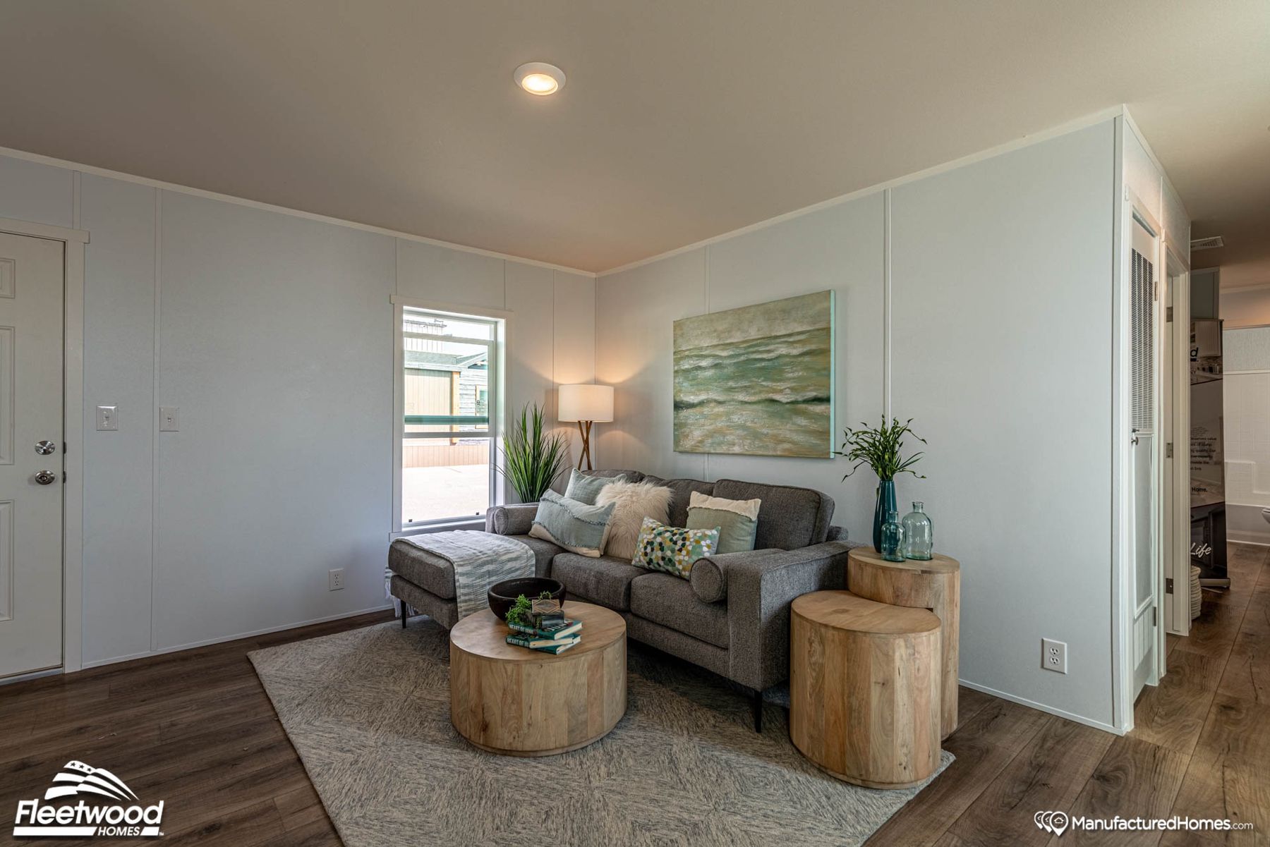 Bright and modern living room with a gray sectional sofa, decorative pillows, round wood coffee tables, and soft natural light from two windows, styled with coastal-themed artwork and plants.