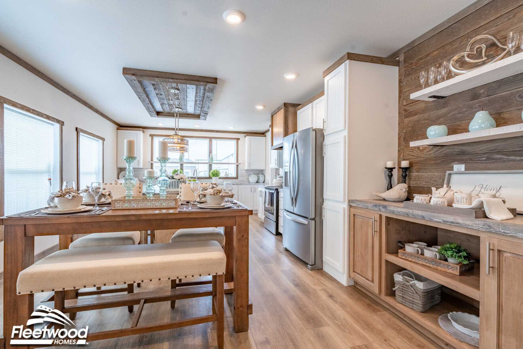 Open-concept kitchen and dining area with a rustic wood island, pendant lighting, wood-look flooring, and large windows, flowing into a living room with a sectional sofa.