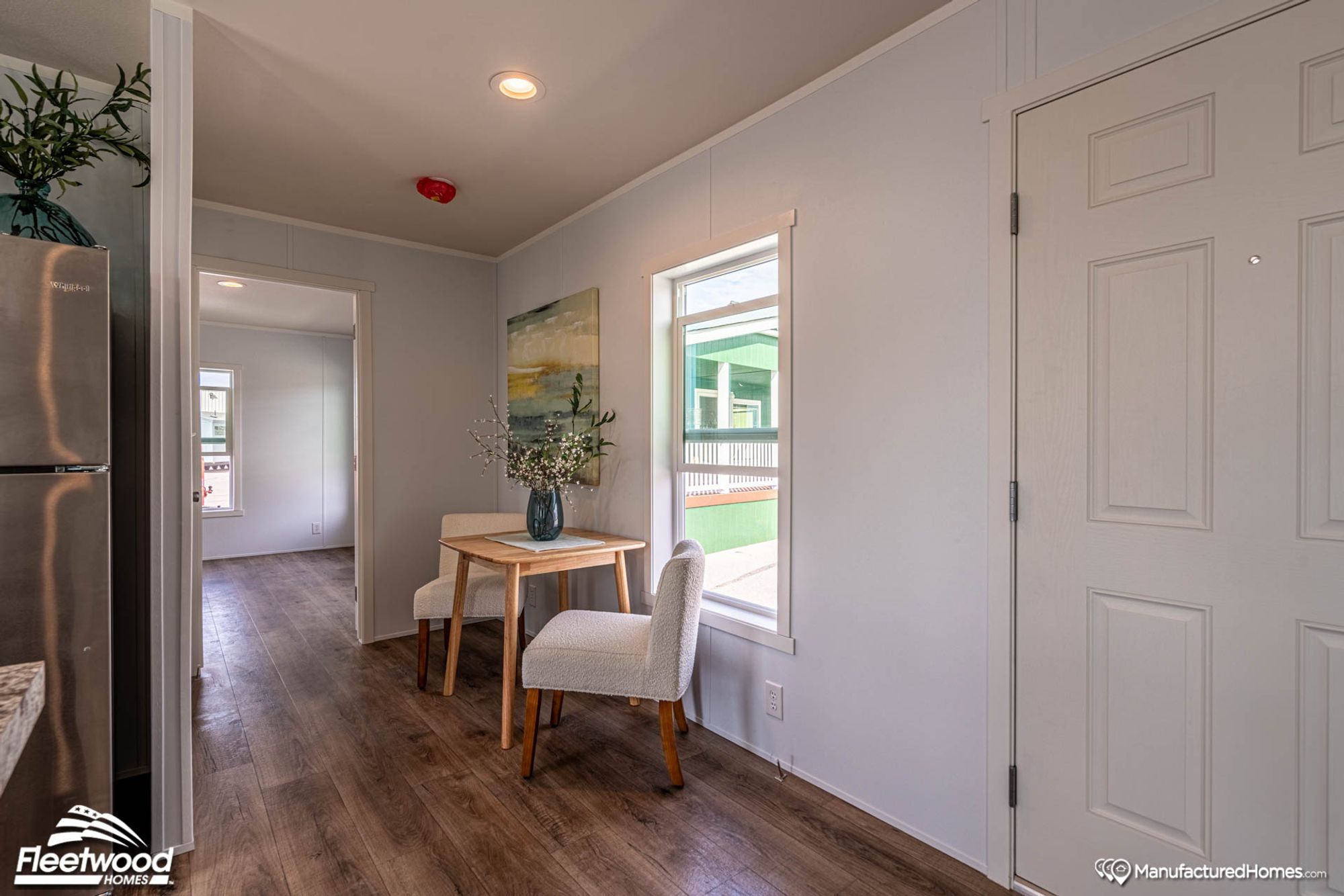 Cozy dining nook with a small wooden table and two upholstered chairs, set beside a window with natural light and decorated with a floral centerpiece and wall art.