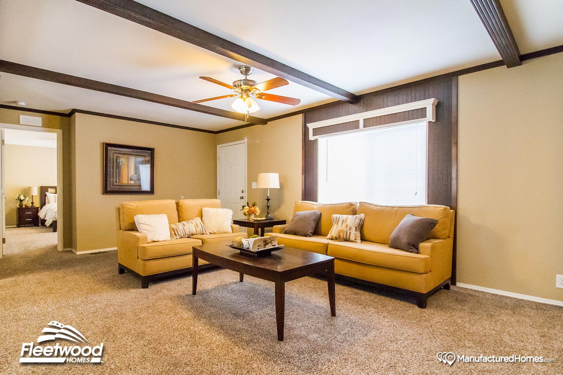 Bright living room in a manufactured home featuring tan sofas with decorative pillows, a dark wood coffee table, carpeted floors, and a ceiling fan with light fixture.