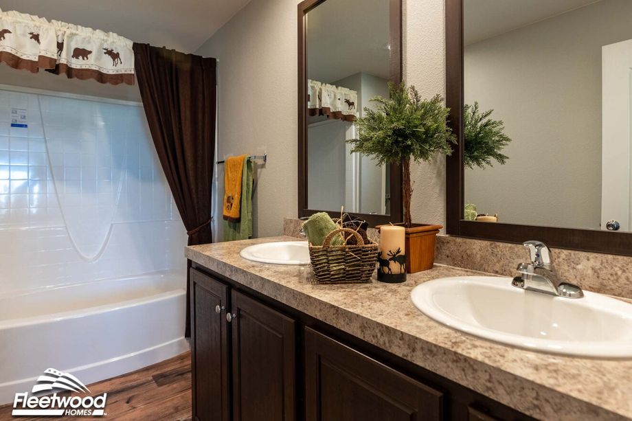 Rustic-themed bathroom with dual-sink vanity, marble countertops, brown and green towels, and a white tub with a moose-print valance and dark shower curtain.