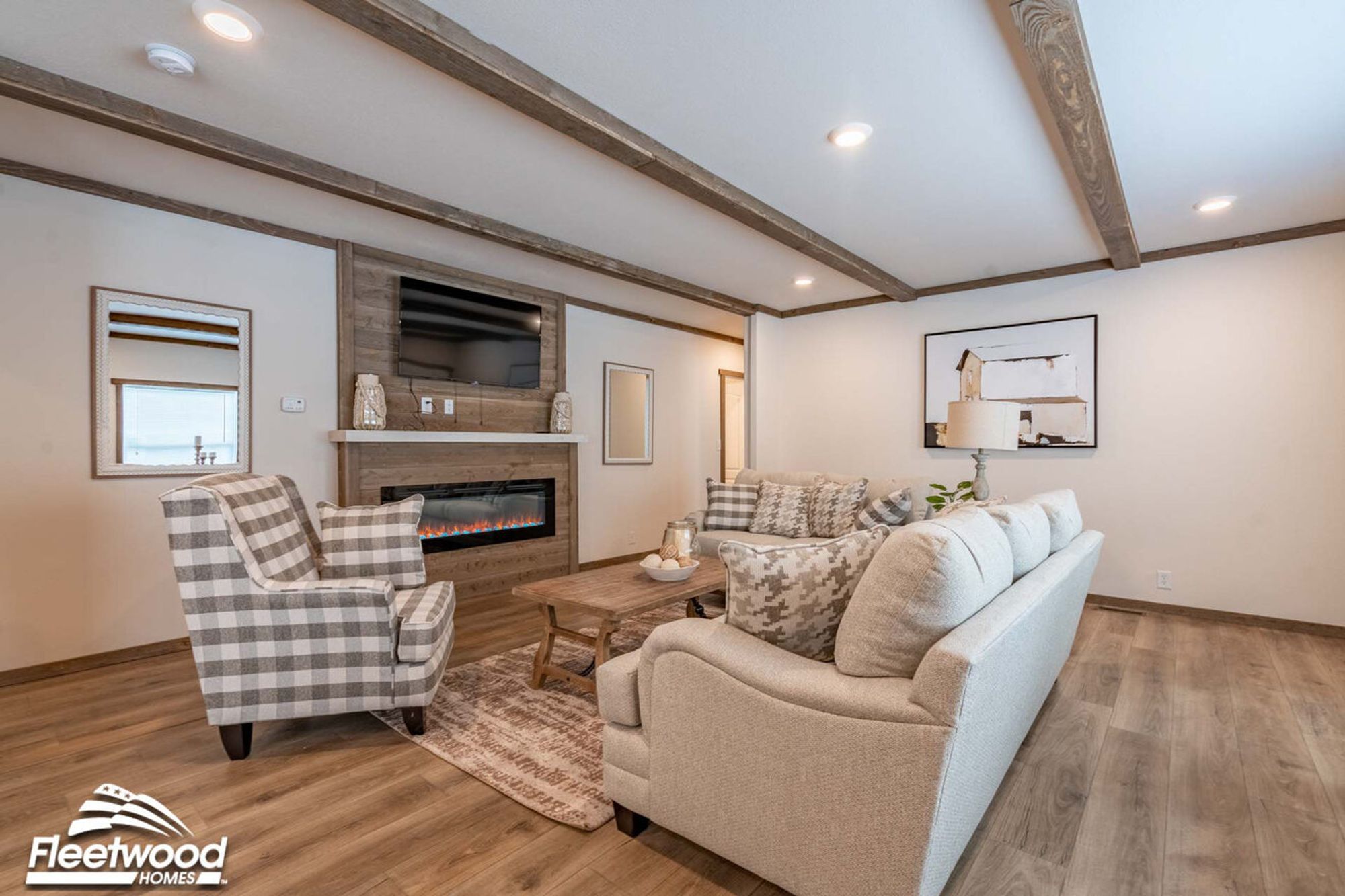 Cozy living room with wood-beam ceiling accents, neutral-toned sofas with patterned pillows, a plaid armchair, a rustic coffee table, and a wood-paneled fireplace wall with a mounted TV.