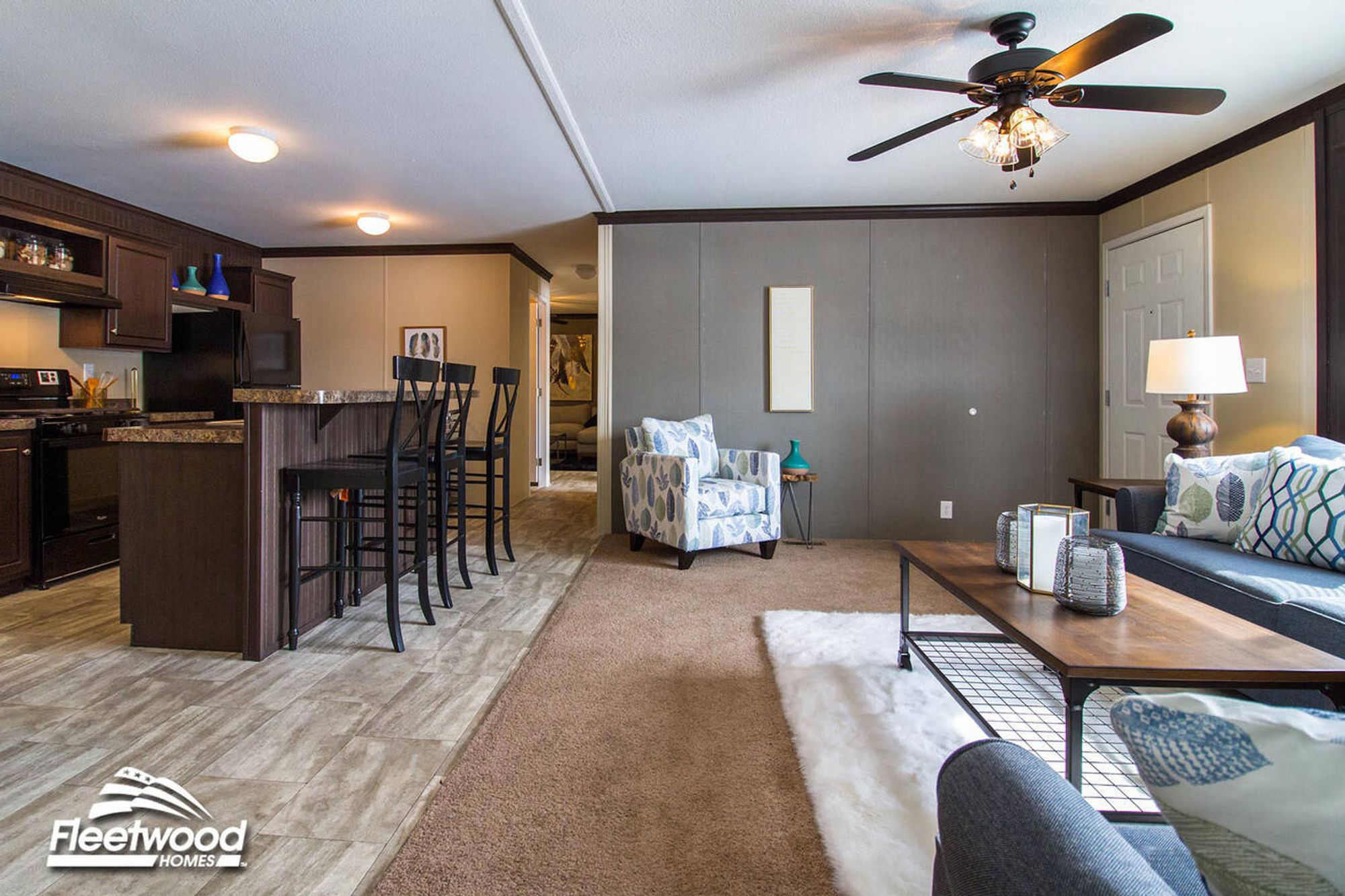 Open-concept kitchen and living room with a breakfast bar and barstools, modern appliances, carpeted seating area with sofas and an accent chair, ceiling fan, and a wooden coffee table on a rug.