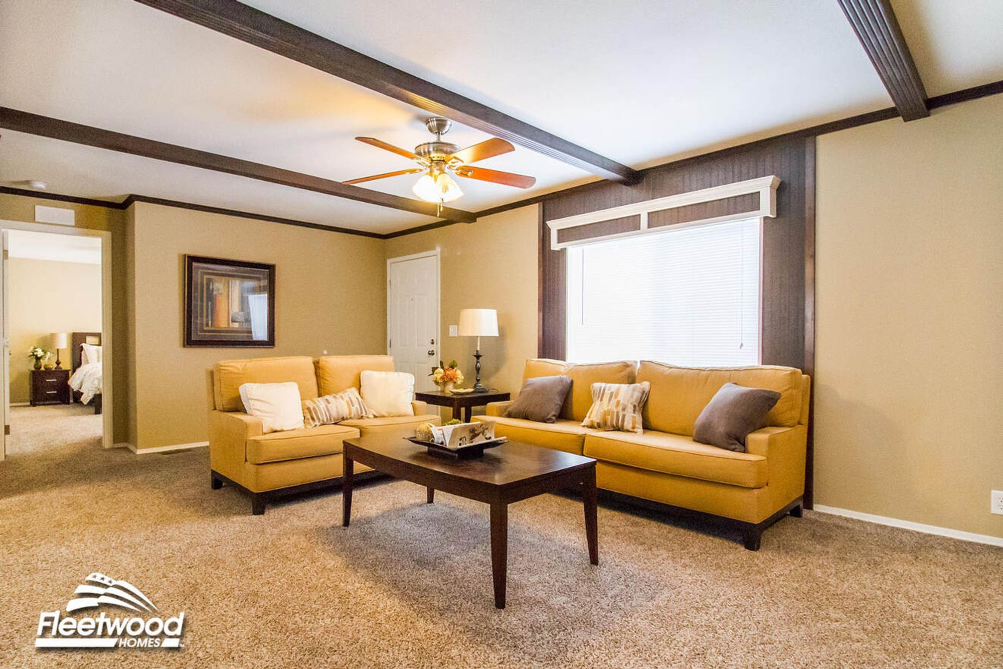 Warm-toned living room with two tan sofas, dark wood coffee table, ceiling fan, and neutral decor, with a bedroom visible in the background.