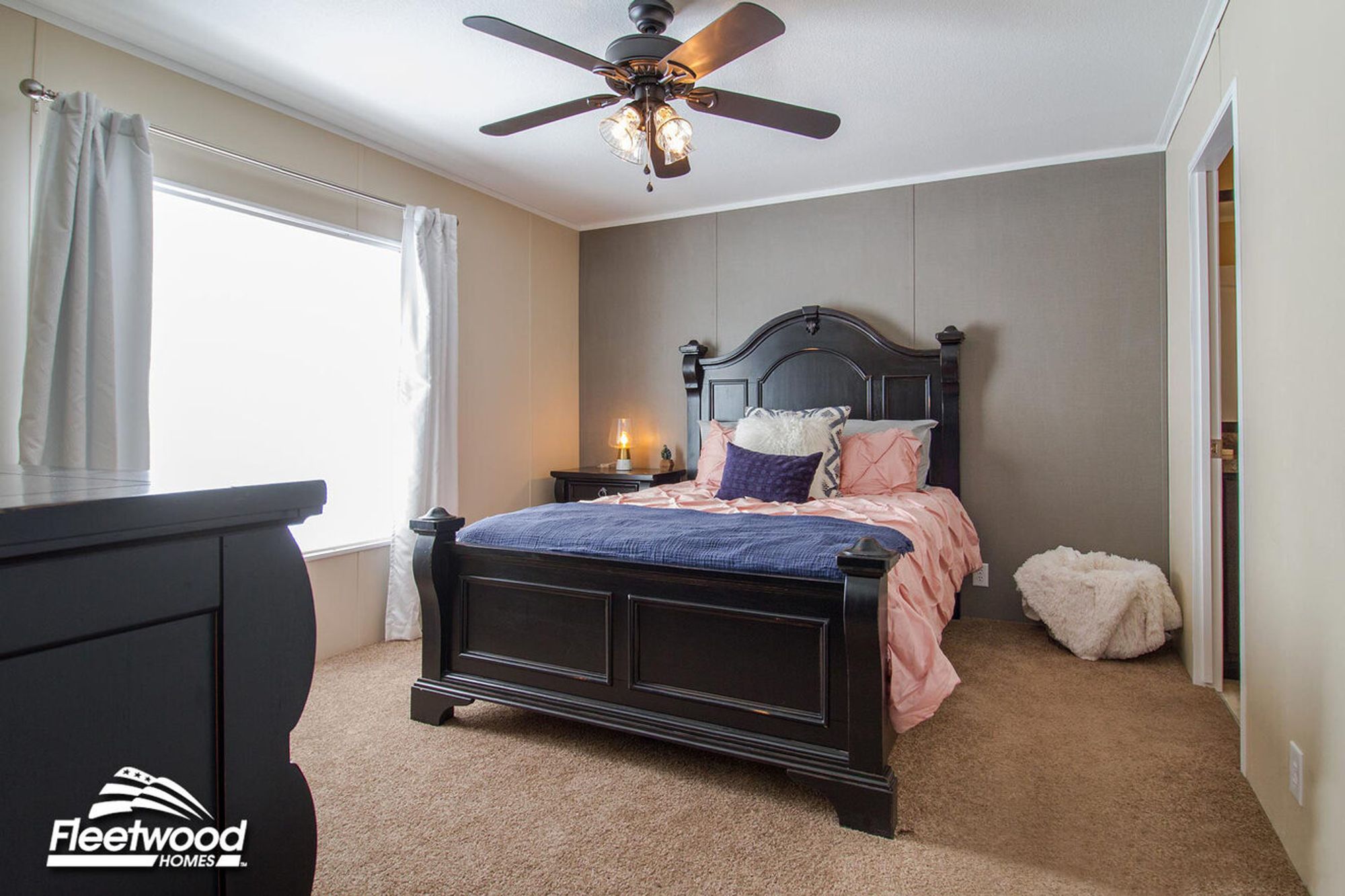 Bedroom with a pink comforter and navy throw blanket on the bed, black dresser with mirror, ceiling fan, and open doors leading to a walk-in closet, bathroom with shower-tub combo, and view into the kitchen.