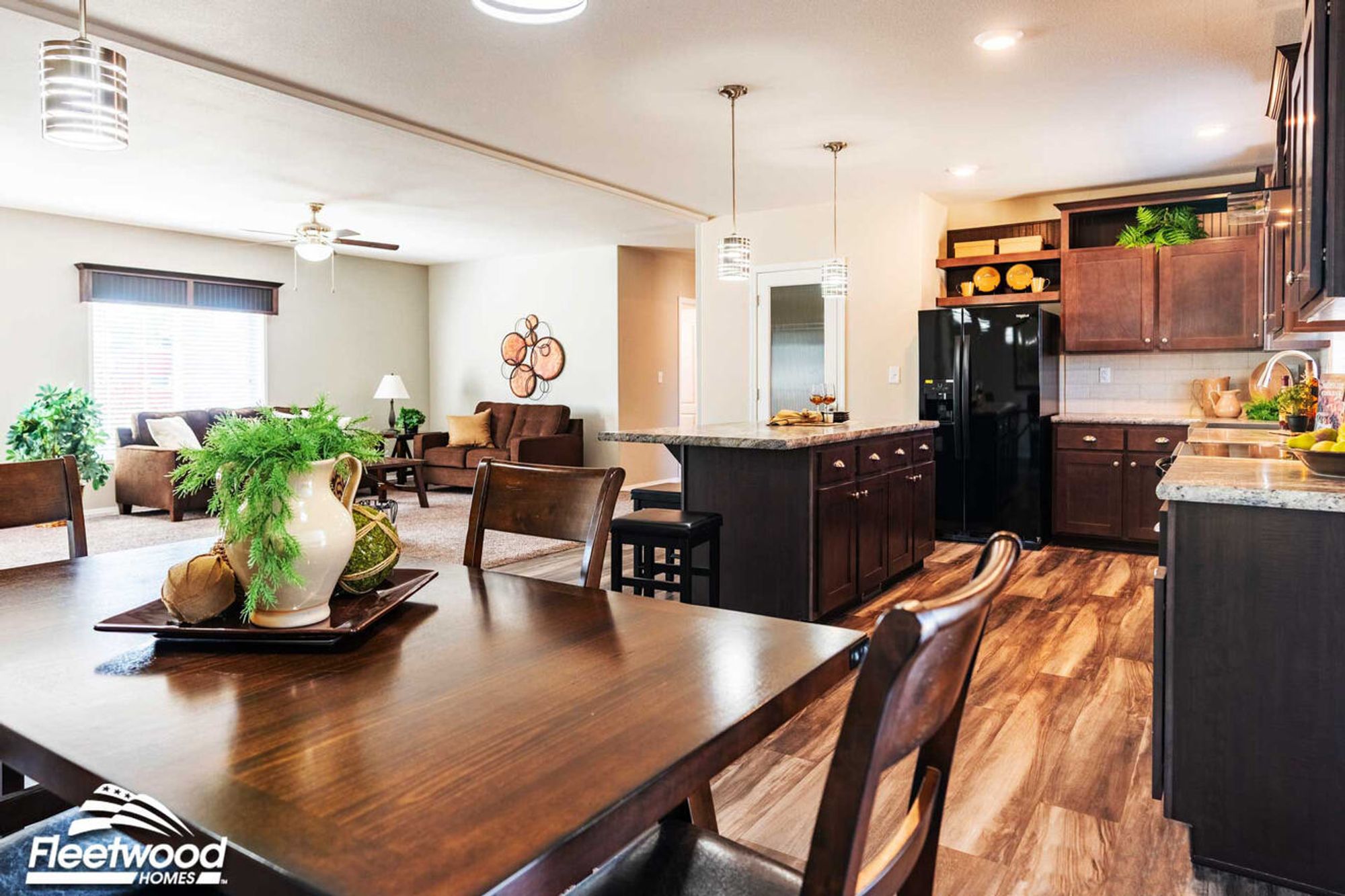 Open-concept dining, kitchen, and living area with dark wood cabinets, center island, modern lighting, and cozy seating by a large window.