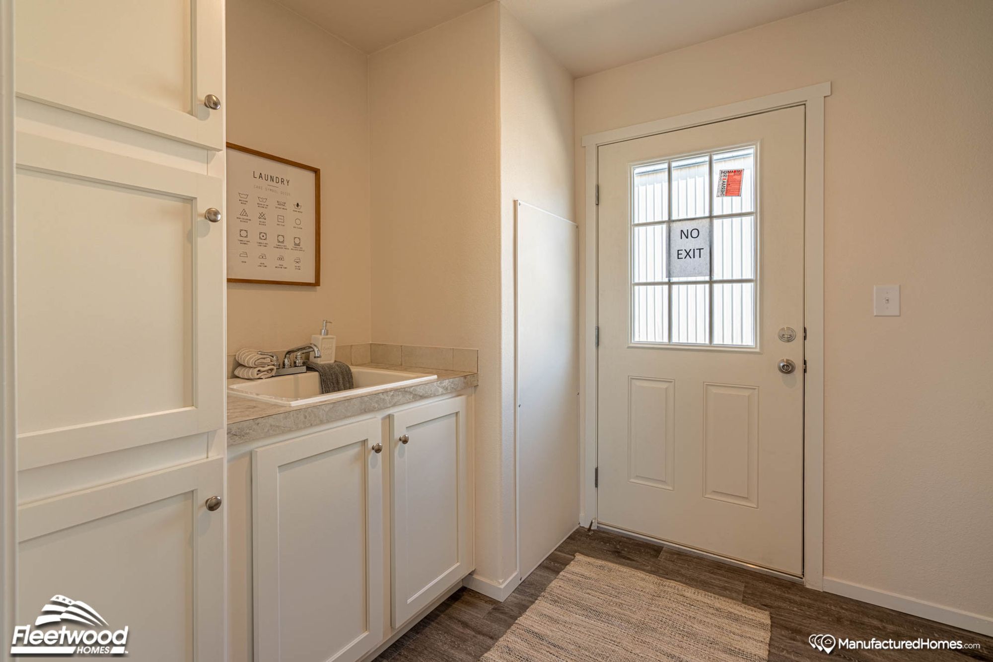Laundry room featuring white cabinetry, a utility sink with marble-style countertop, and a door marked "No Exit" with window panels. The space includes a decorative laundry guide and folded towels for a clean, organized look.
