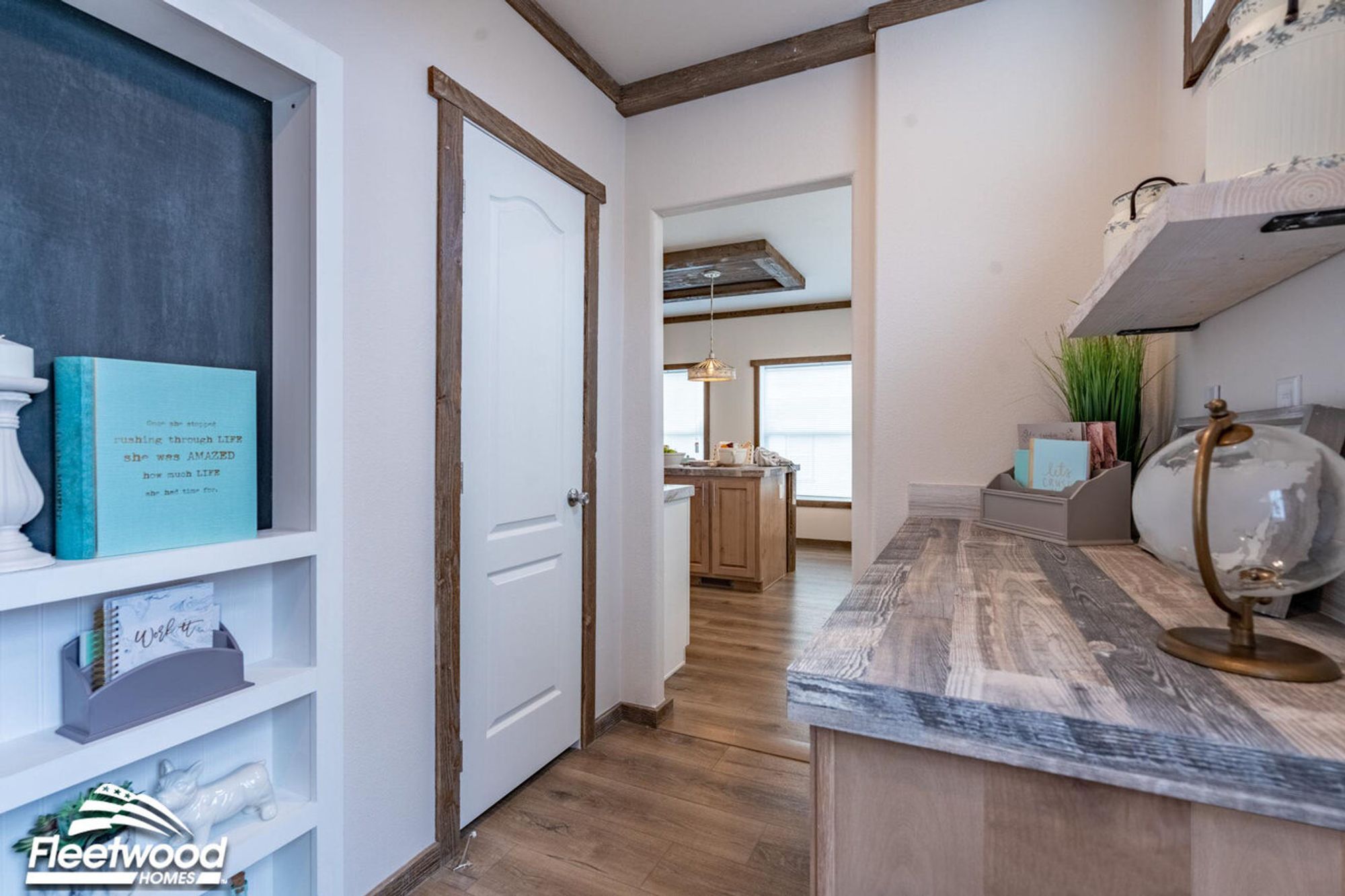 Hallway with rustic wood accents featuring a built-in desk with decorative items, shelves with books and decor, and a view into a bright kitchen with pendant lighting.