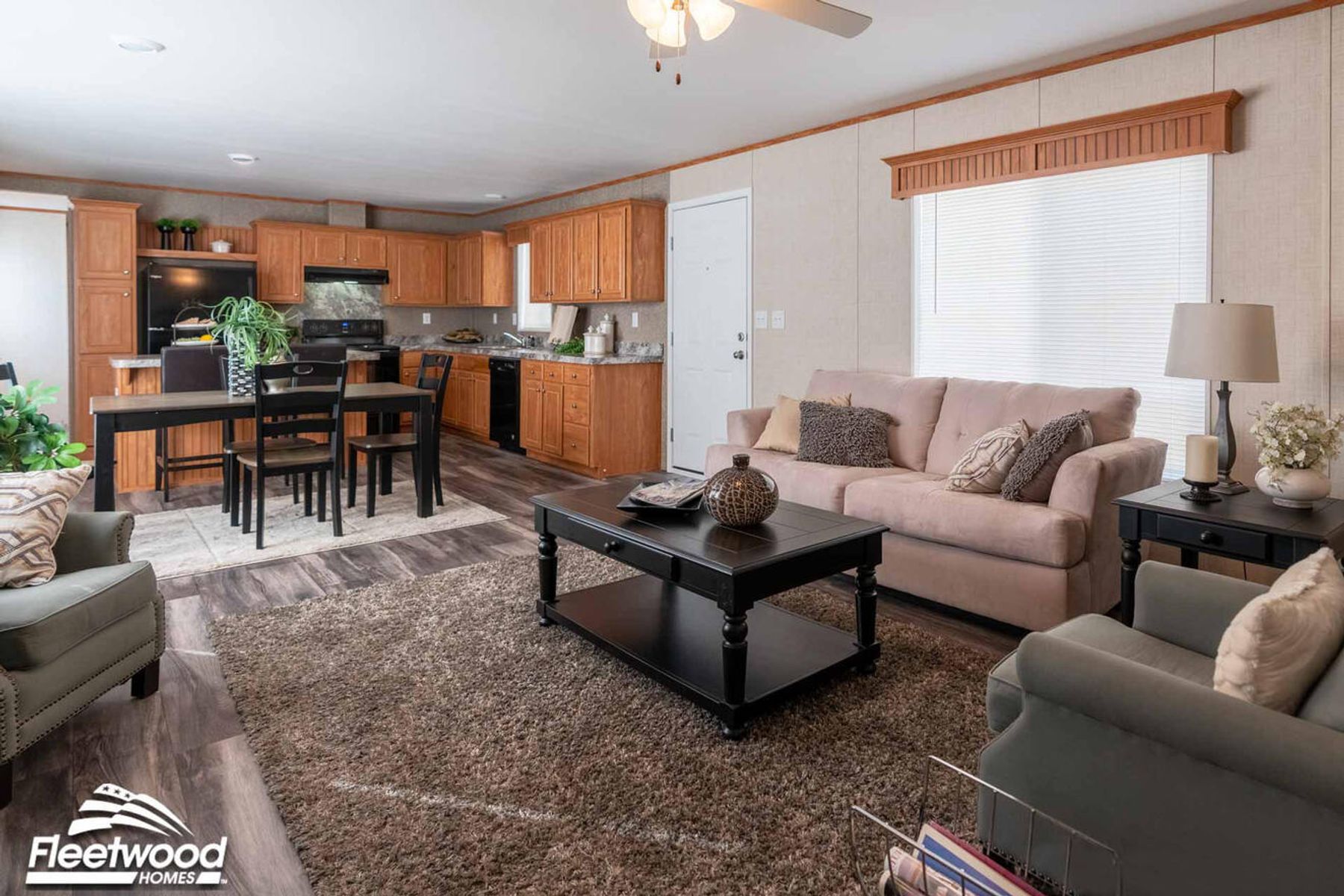 Living and dining area with dark wood furniture, a black cabinet, armchair, and coffee table, decorated with plants and wall art, opening into a kitchen with wood cabinets and black appliances.