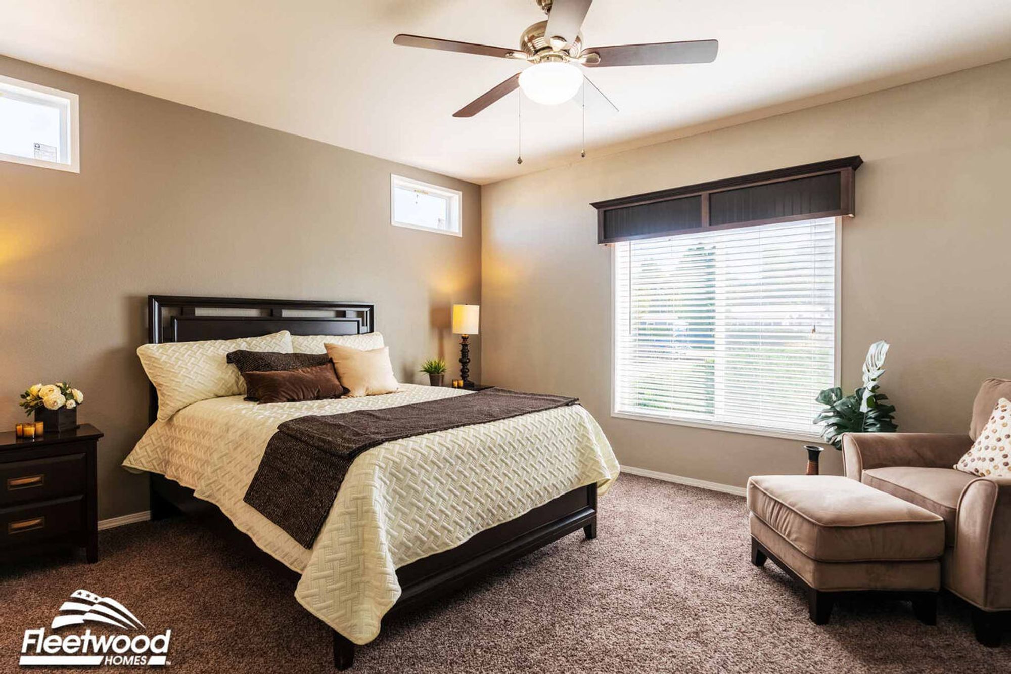 Bedroom with neutral decor, featuring a bed with white and brown bedding, dark wood dresser, beige armchair, and ceiling fan.