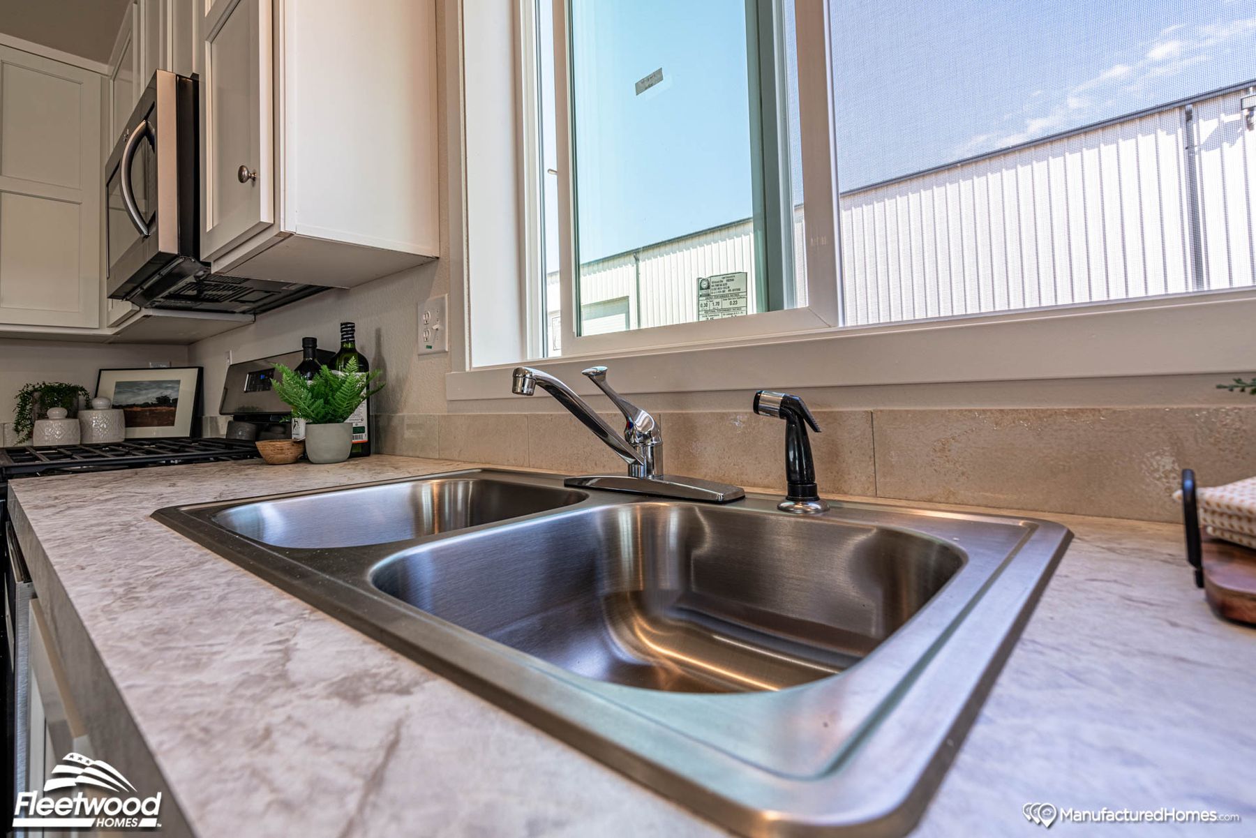 Close-up of a double-basin stainless steel kitchen sink with a chrome faucet and side sprayer, set in a marble-style countertop. A large window above the sink provides natural light, with a tiled backsplash and minimal decorative items nearby, including bottles and a small potted plant.
