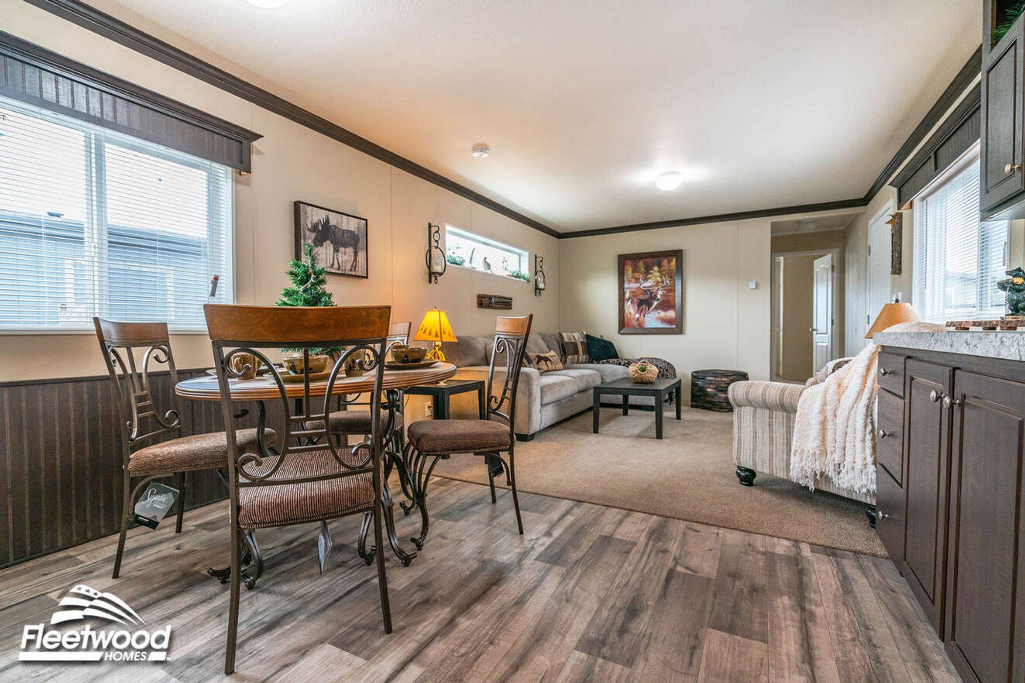 Open dining and living area with a round dining table, cushioned chairs, a sectional sofa, and a coffee table, decorated with rustic wall art.