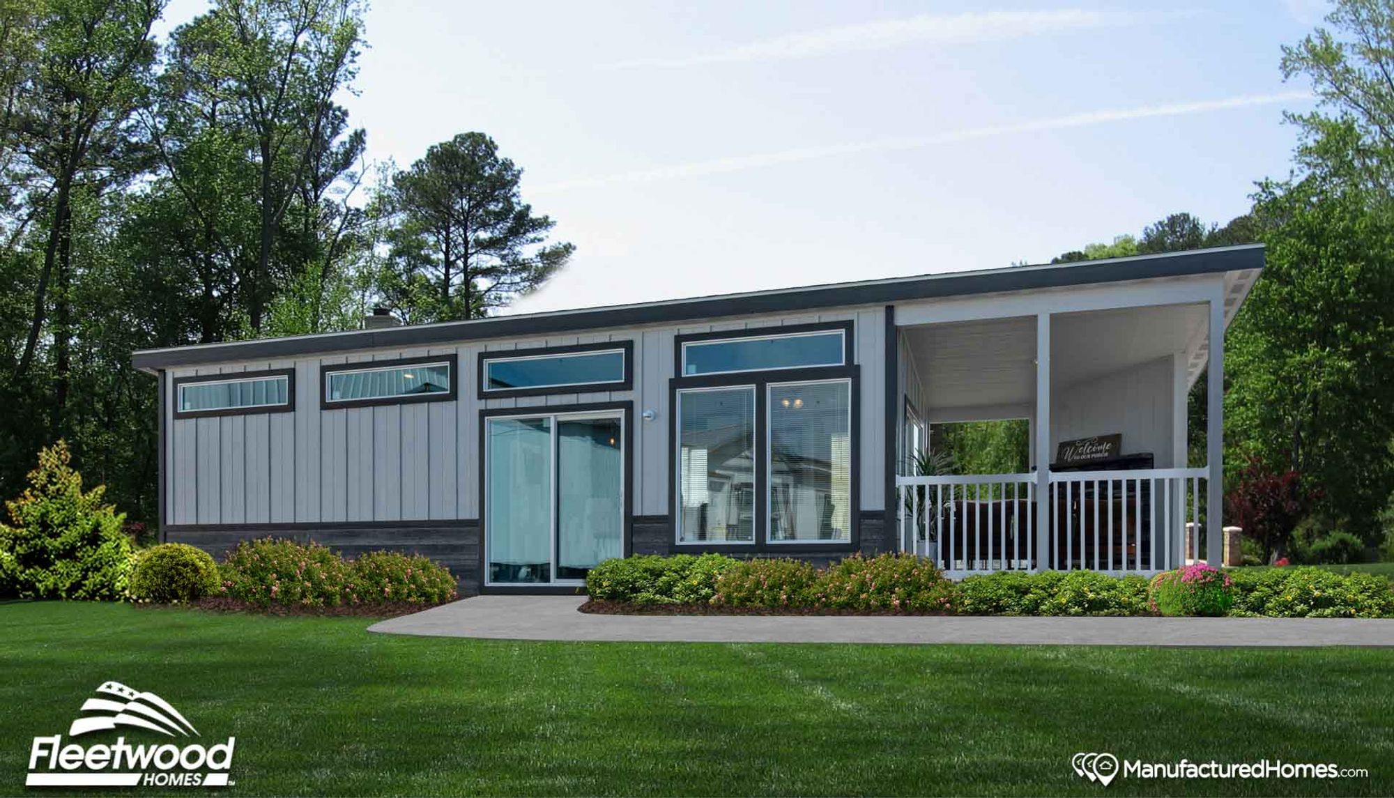 Modern manufactured home with gray siding, large windows, sliding glass doors, and a covered front porch with white railing, surrounded by green lawn and landscaped shrubs.