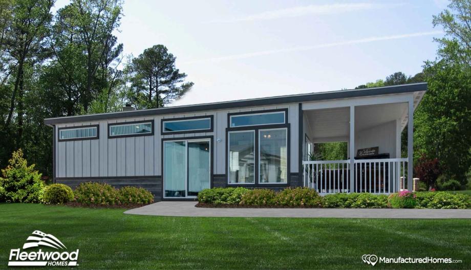 Modern manufactured home with gray siding, large windows, sliding glass doors, and a covered front porch with white railing, surrounded by green lawn and landscaped shrubs.