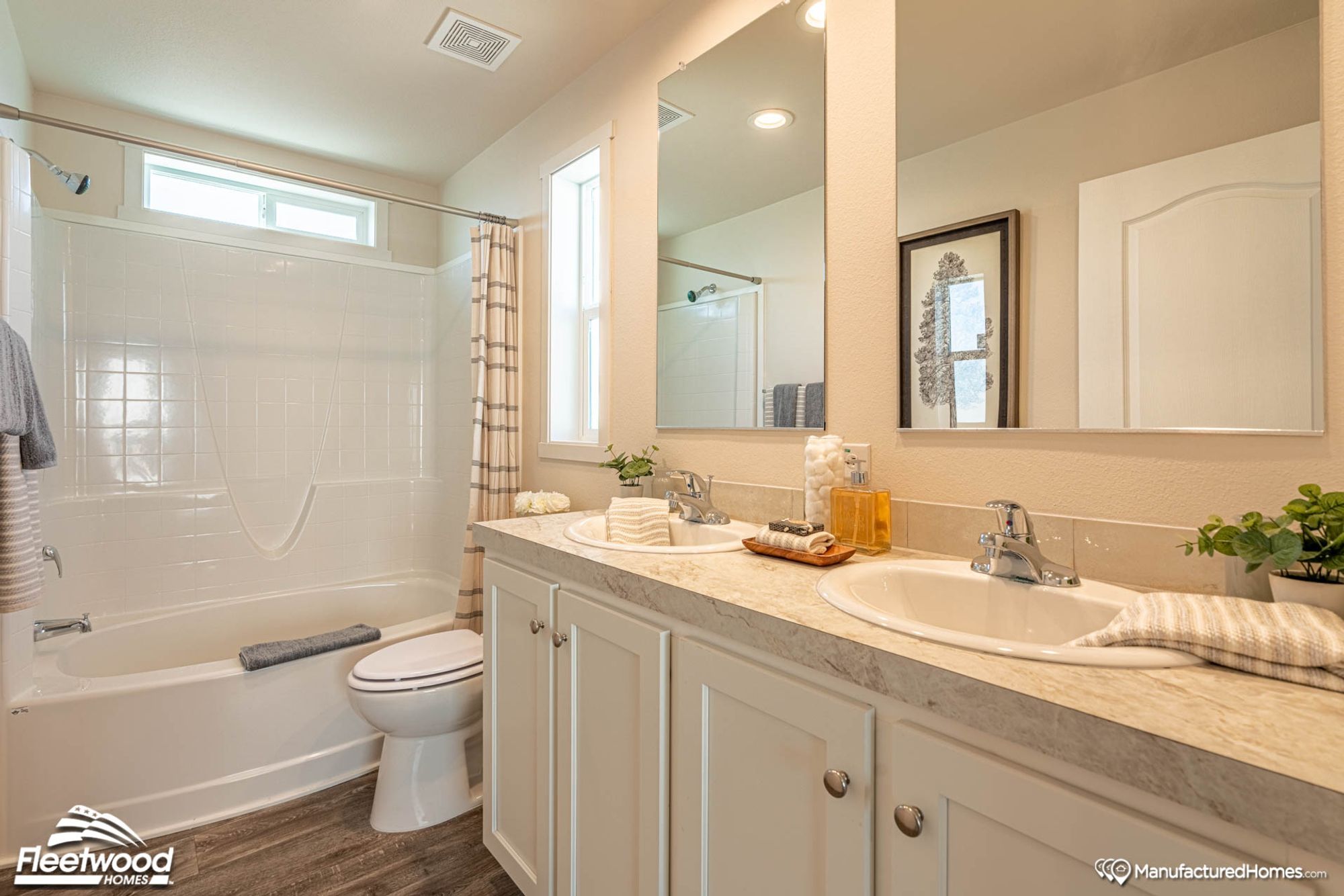 Bright bathroom with a white shower-tub combo, striped shower curtain, and window above. Gray and striped towels hang nearby, with white flowers placed on the toilet tank lid.