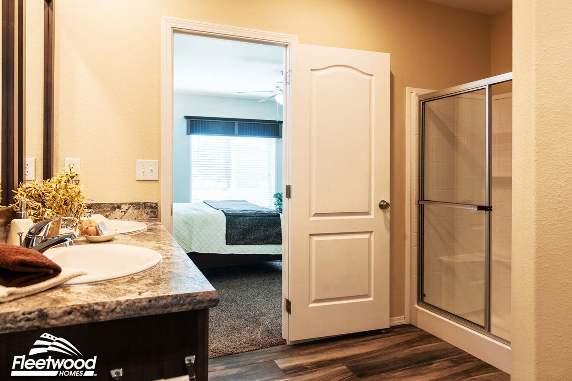 Bathroom with double sinks, granite countertop, and glass-enclosed shower, leading into a bedroom with natural light.