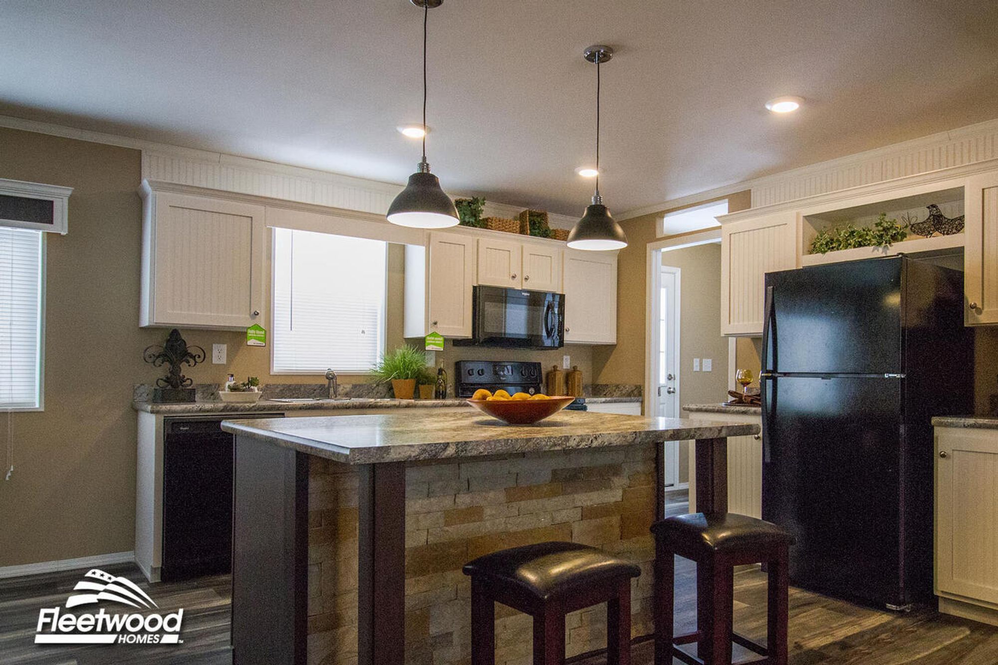 Modern kitchen with white cabinetry, stone island, black appliances, and pendant lighting, featuring barstools and decorative greenery.