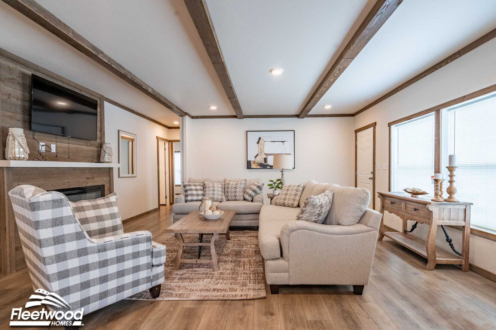 Cozy living room with wood-beam ceiling accents, neutral-toned sofas with patterned pillows, a plaid armchair, a rustic coffee table, and a wood-paneled fireplace wall with a mounted TV.